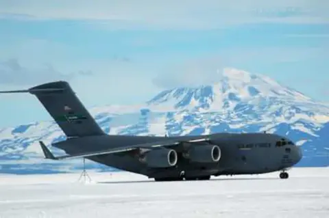 A large military jet transport parked on a snowy ramp with a volcano behind it