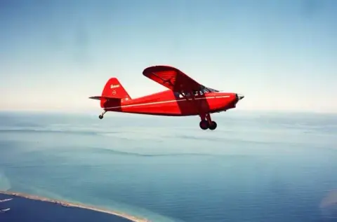 A red high-wing taildragger in flight over water