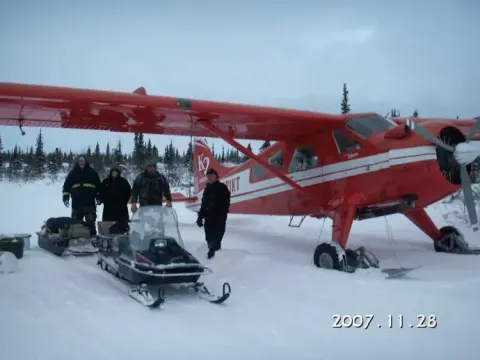 A red ski-equipped taildragger parked on snow with snowmobiles nearby