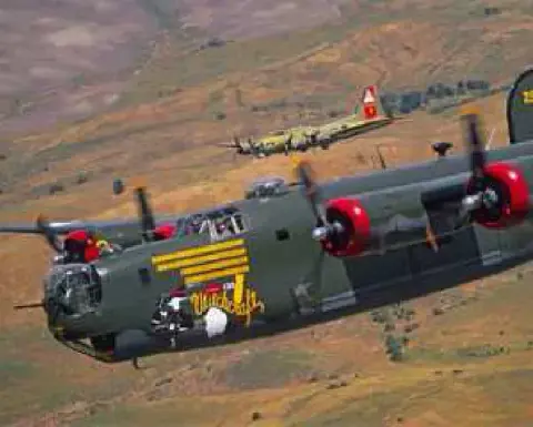 A camouflaged B-24 bomber with yellow tail stripes in flight over hills, another bomber behind