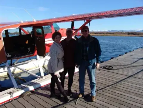 Three people standing on a dock in front of a red floatplane