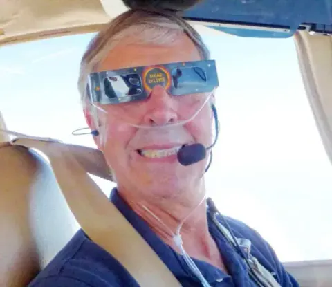 A pilot wearing eclipse glasses and a headset in the cockpit