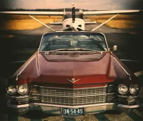 A vintage red Cadillac convertible with a small airplane behind it