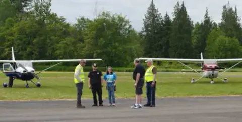 Volunteers in safety vests on the ramp with small airplanes behind