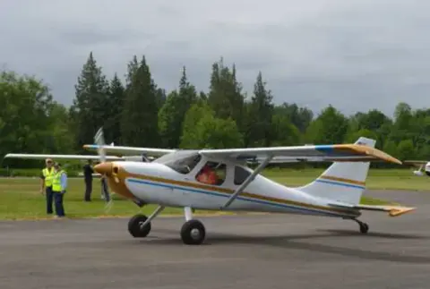 A white high-wing airplane with gold trim taxiing past volunteers