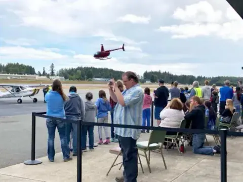 Spectators along a fence watching a red helicopter take off