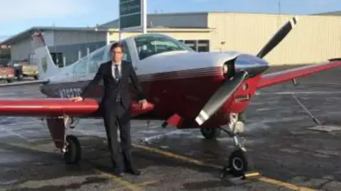 A young man in a suit standing beside a red and white low-wing airplane on the ramp