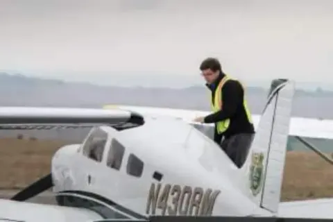 A young man in a safety vest beside the tail of a white low-wing airplane
