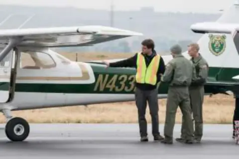 Three men in safety vests and uniforms beside a white high-wing airplane