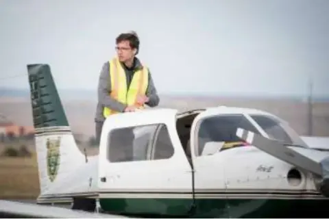 A young man in a safety vest standing on the wing of a white low-wing airplane