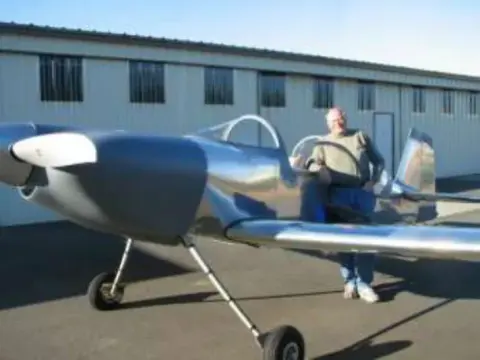 A man in the cockpit of a polished metal low-wing airplane outside a hangar