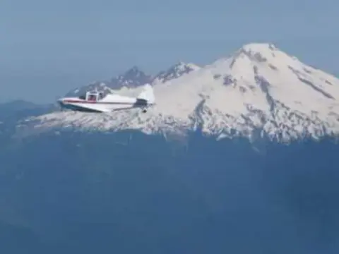 A small white airplane flying past a snow-covered volcano