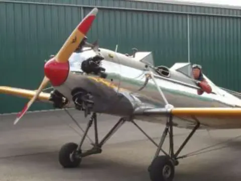 A silver and yellow open-cockpit airplane with a wooden propeller in front of a hangar