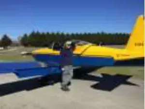 A man in a jacket standing beside a yellow and blue low-wing airplane on the grass