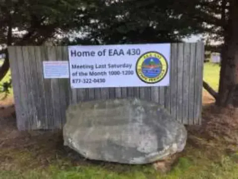 A Home of EAA 430 sign on a wooden fence above a large boulder