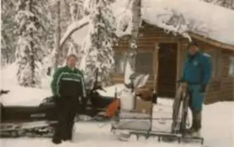 Two men with a snow machine and a loaded sled outside a snowy log cabin