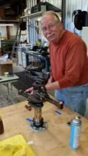 A smiling man in a red shirt working at a bench with a part in a vise