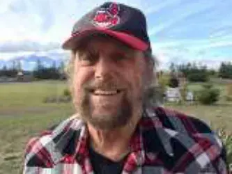 A smiling bearded man in a red and black cap and plaid shirt outdoors