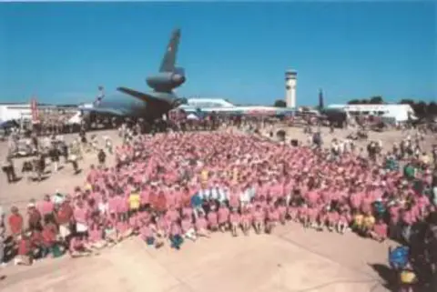 A large crowd in pink shirts posed in front of a vintage airplane at an airshow