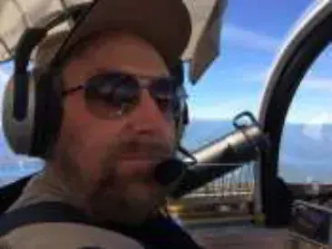 A smiling bearded pilot in a cap, sunglasses and headset