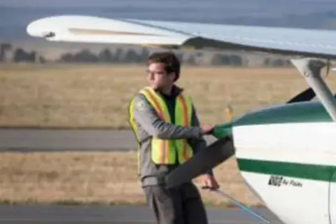 A young man in a safety vest beside the wing of a parked airplane