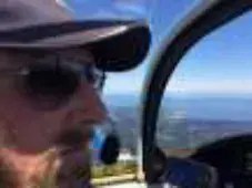 A smiling bearded pilot in a cap, sunglasses and headset