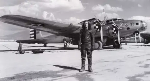 A black and white photo of a man standing by a World War II era airplane