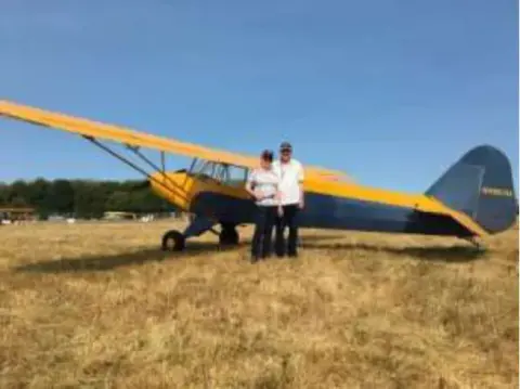 A couple standing beside a yellow and blue high-wing airplane on the grass