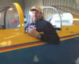 A smiling bearded man in a cap in the cockpit of a yellow airplane