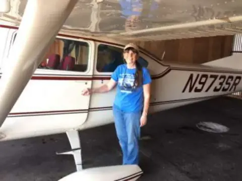 A woman standing beside the door of a white high-wing airplane in a hangar