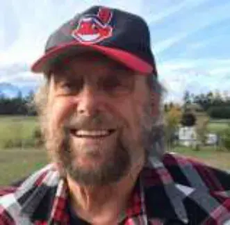 A smiling bearded man in a red and black cap and plaid shirt outdoors