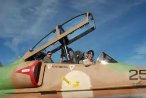 Two pilots in the open cockpit of a camouflaged jet trainer
