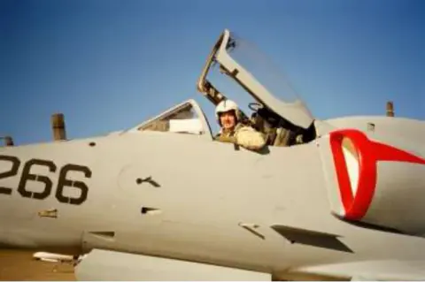 A pilot waving from the open cockpit of a gray jet numbered 266