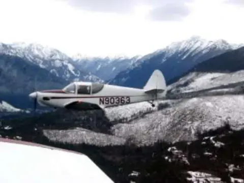 A white high-wing airplane in flight over snowy mountains