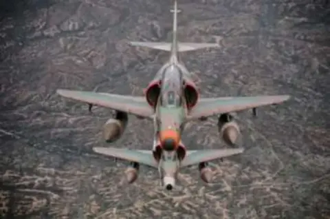 A camouflaged Skyhawk jet with drop tanks in flight, seen from above