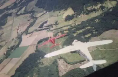 A white biplane and a red airplane flying over farmland, seen from above