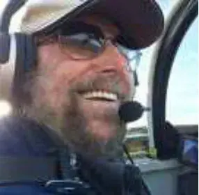 A smiling bearded pilot in a cap, sunglasses and headset