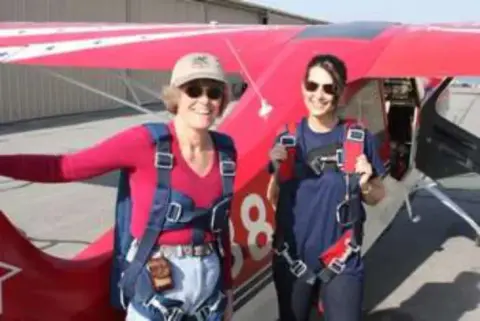 Two women in parachute harnesses in front of a red aerobatic airplane