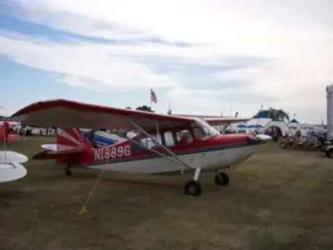 A red and white high-wing airplane parked on grass at a fly-in