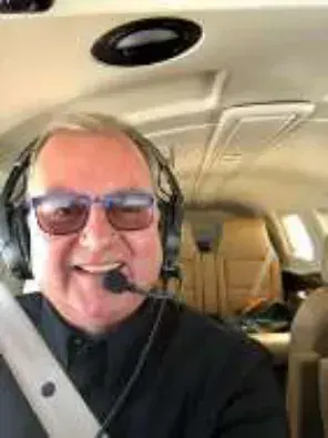 A smiling white-haired man in sunglasses and a headset in a cockpit