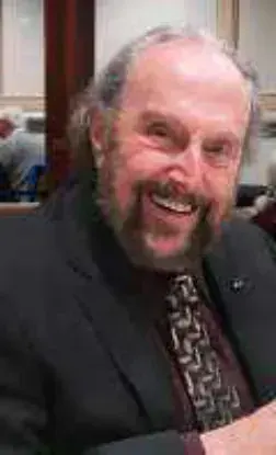 A smiling bald man with a white beard in a dark jacket and patterned tie