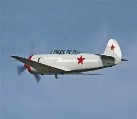 A silver Yak trainer with a red star in flight