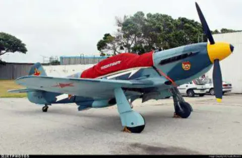 A silver and red Yak fighter with a large propeller parked on the ramp