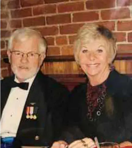 A white-haired man in a tuxedo with medals and a smiling blonde woman, seated in front of a brick wall