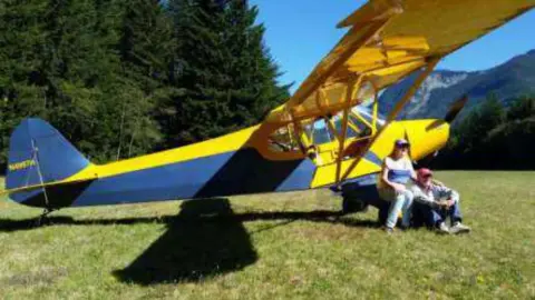 A yellow and blue Cub-style airplane parked on grass with people beside it