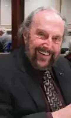 A smiling bald man with a white beard in a dark jacket and patterned tie