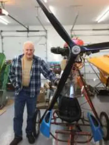 A white-haired man in a plaid shirt beside a propeller and an airplane under construction in a hangar