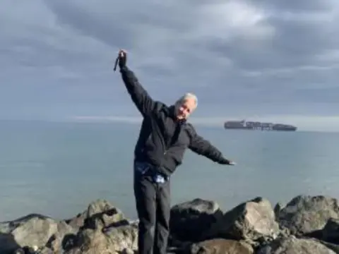 A man posing with arms raised on rocks by the water