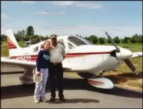 A man and a woman standing next to a red and white low-wing airplane