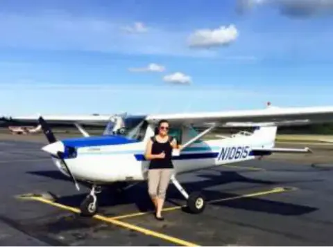 A woman standing beside a white and blue high-wing airplane on the ramp